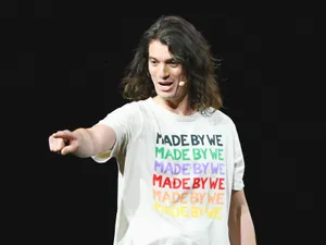 Adam Neumann, standing on stage wearing a microphone and a white shirt that says "Made by We" repeatedly in rainbow colors, pointing at the audience