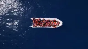 Still frame of an inflatable boat full of people wearing orange life jackets, pictured from above
