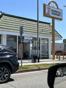 A photograph of a restaurant with a sign featuring a Bored Ape NFT and the text "Bored & Hungry". The building is vacant and a sign shows "Chile Verde Mexican Food coming soon"