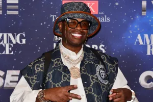A man wearing a denim bucket hat and vest with the Gucci logo, a white button-down shirt, large black glasses, a massive gold and diamond chain, and two watches, poses in front of a background