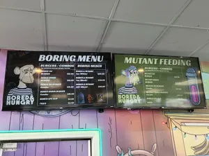 Two television screens showing a beef menu and vegan menu, emblazoned with Bored Apes and Mutant Apes. Prices start at $13 for beef hamburgers and $15.50 for vegan burgers.