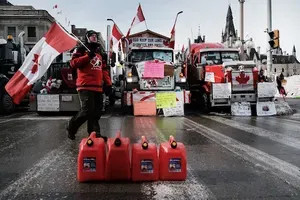 18-wheeler trucks plastered in signage, with a man walking in front waving a Canadian flag. There are several plastic fuel canisters on the ground in the foreground.
