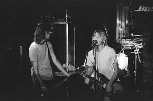 A black and white photo of Cobain singing and playing guitar, with another guitarist next to him