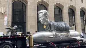 A large silver statue of Elon Musk's head, atop a rocket shaped structure. The sculpture is on the back of a flatbed truck.