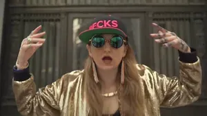 A woman in round sunglasses wearing a shiny gold jacket and a baseball cap that says "#0FCKS" sings with her hands in the air