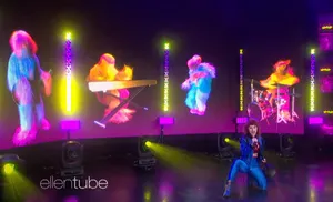 Four figures resembling neon-colored versions of bigfoot play instruments on a large screen. A woman wearing bright blue pants and a jacket kneels in front of the screen singing into a microphone.