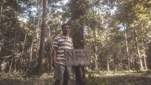 Aman in a polo shirt stands in the rainforest with a sign reading "NFT"